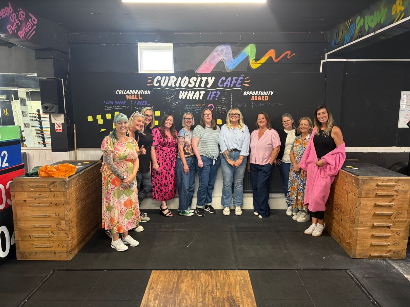 Twelve women standing together in front of the Curiosity Caf&eacute; collaboration wall at Weights and Cakes, covered in handwritten notes and sticky notes