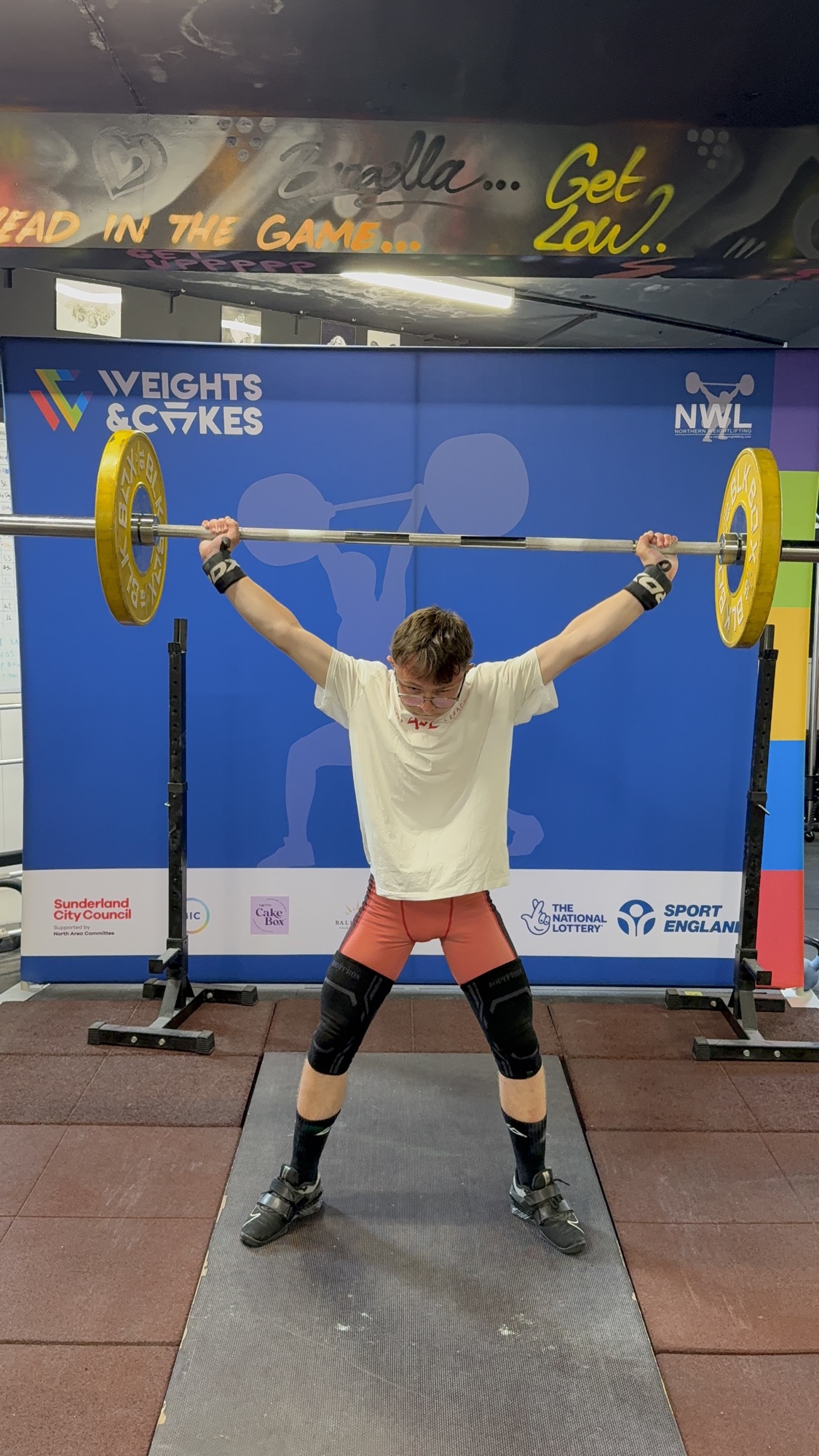 A young lifter holding a barbell overhead in front of the club competition backdrop